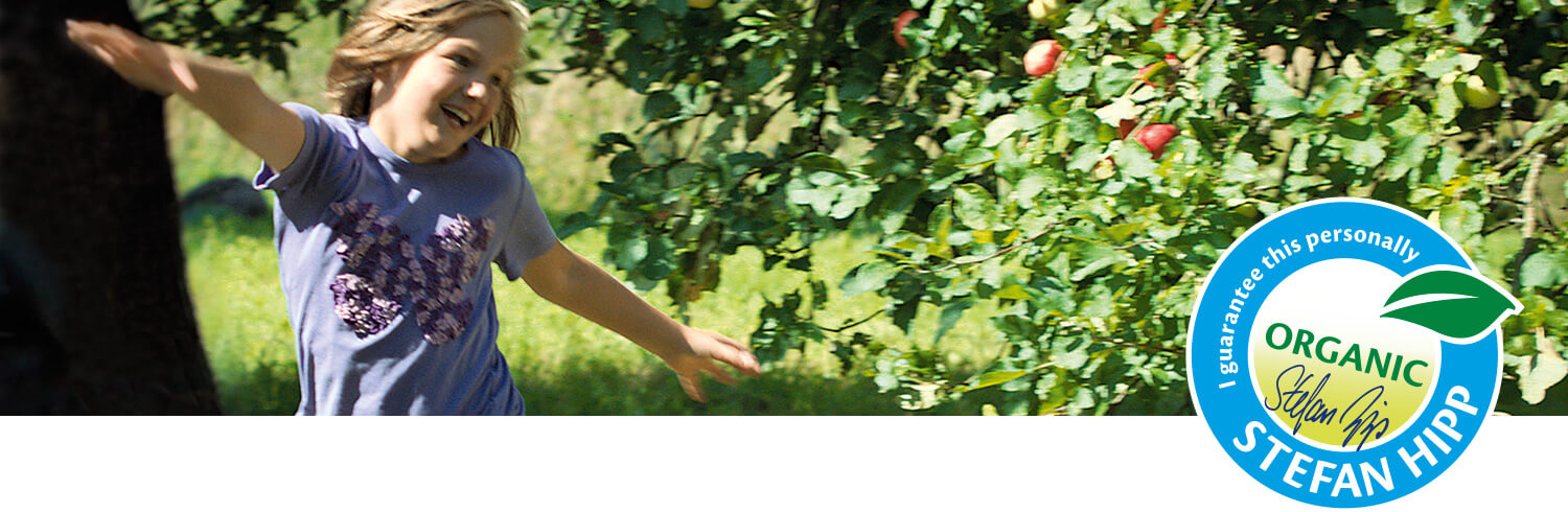 A child in a garden with a tree bearing fruit, wearing a purple shirt with a floral design, is captured in a candid moment.