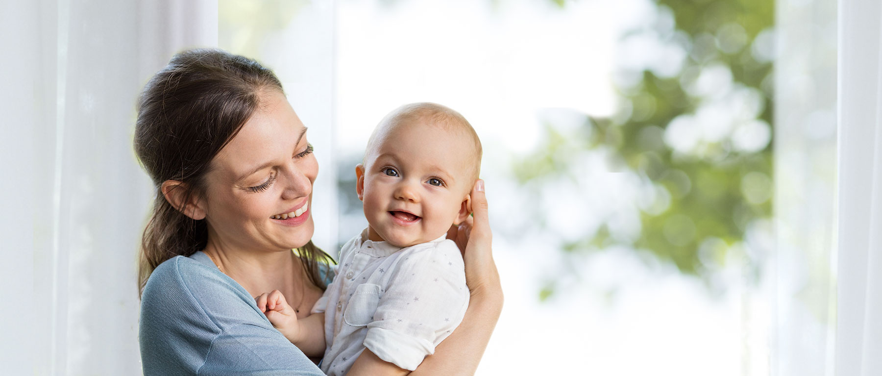 A woman in a blue shirt holds a baby in a white outfit, both looking towards the camera, with a blurred background of greenery and a window.