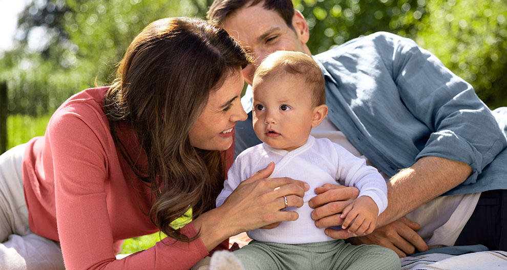 A family of three, consisting of a man, a woman, and a baby, are engaged in a playful interaction outdoors.