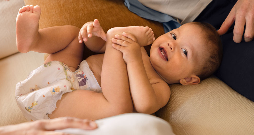 A baby, wearing a diaper with colorful illustrations, is lying on a couch, surrounded by adults' hands, with one hand gently touching the baby's foot.