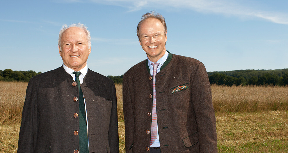 Two men in suits stand outdoors against a backdrop of a field and trees.