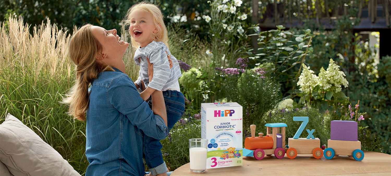 Naine ja laps naeravad koos õues, klaas piima ja karp Hipp Junior Combiotic'u kõrval mängurongiga.