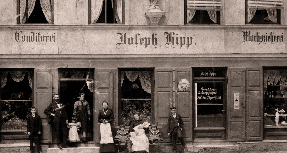Grupp inimesi, sealhulgas täiskasvanud ja lapsed, seisavad ja istuvad poe ees, millel on silt "Johann Hipp".